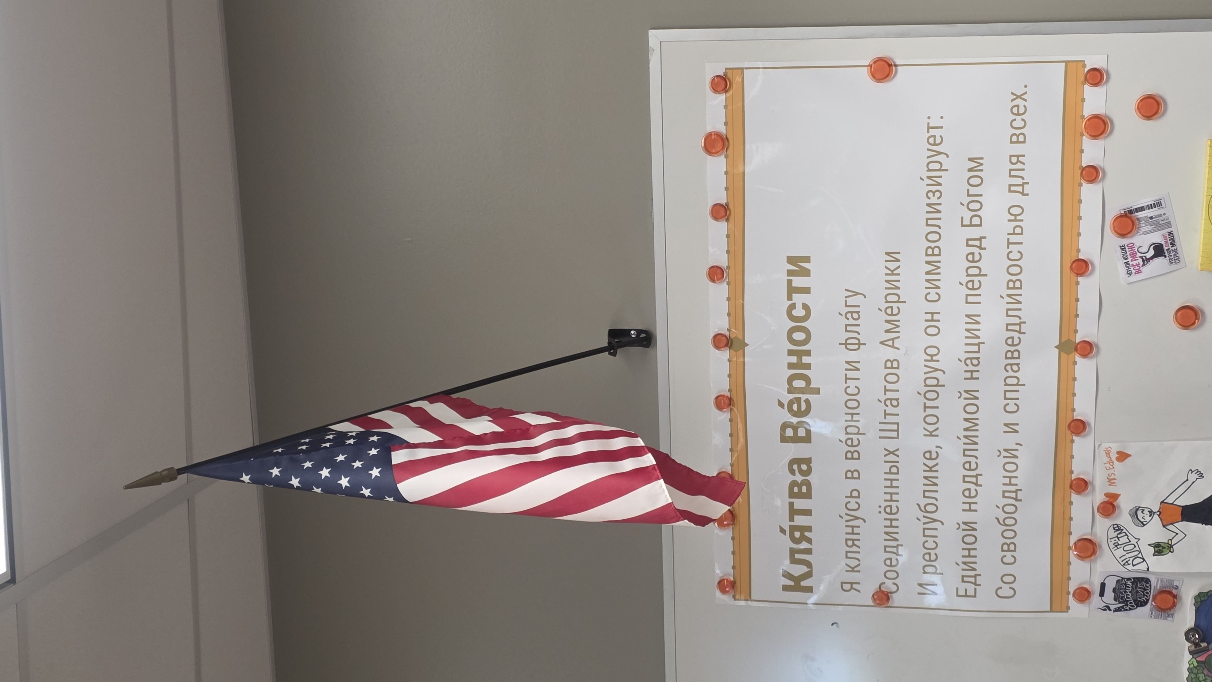 A classroom American flag; underneath is a poster with the words of The Pledge of Allegiance in Russian